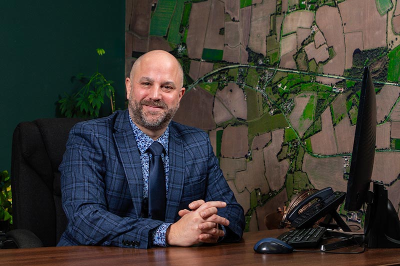Man in a blue plaid suit sits at a desk with his hands clasped, smiling. Aerial map on the wall behind and a computer monitor on the desk.