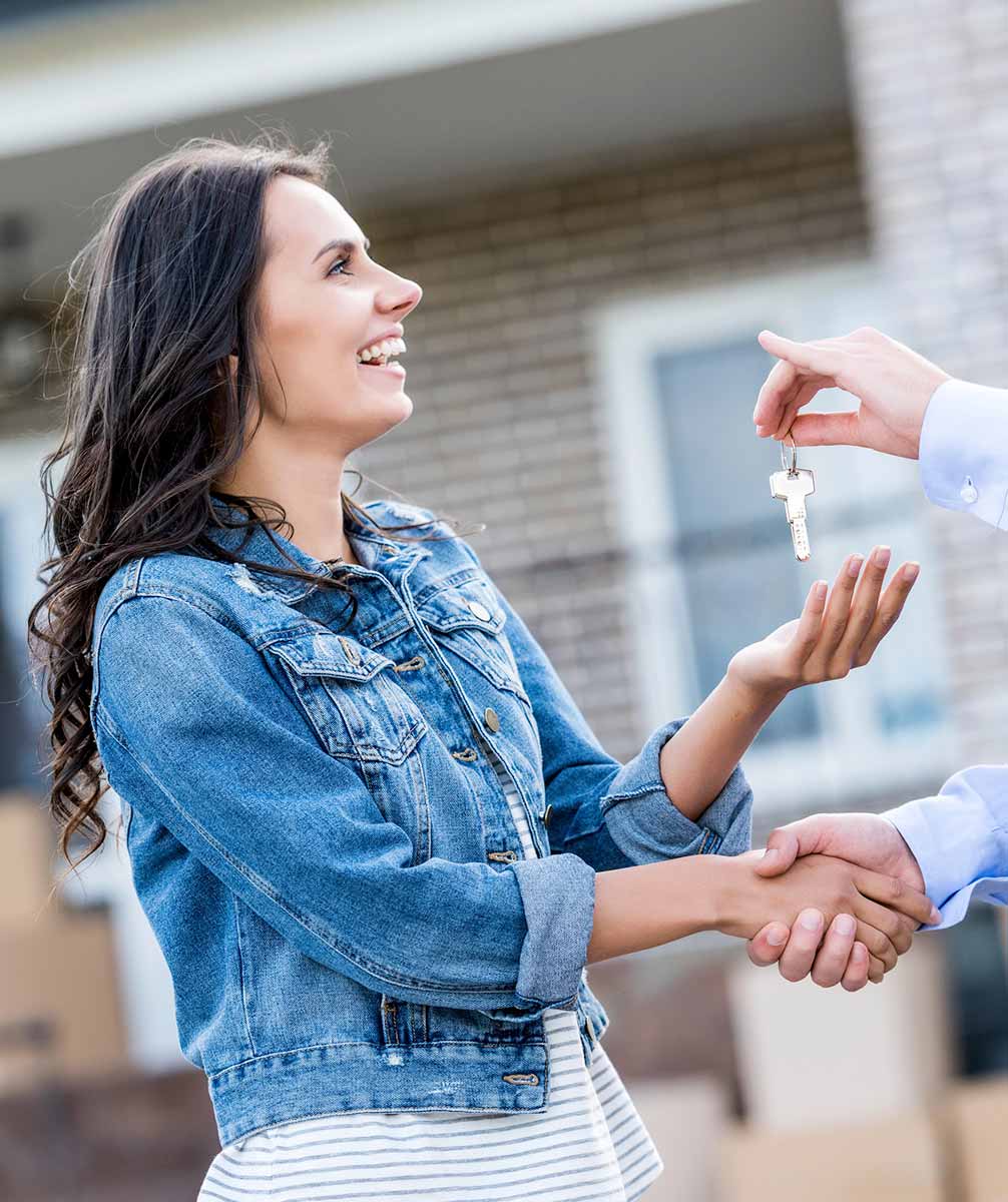 A woman in a denim jacket smiles as she receives a key from another person, shaking hands.