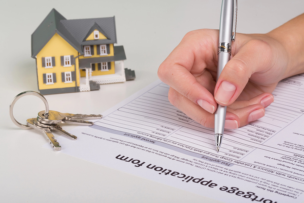 A hand signs a mortgage application form with a pen, poised for selling your home in Dereham. Beside the document sits a model house and a keyring with keys, symbolizing the start of new ownership.