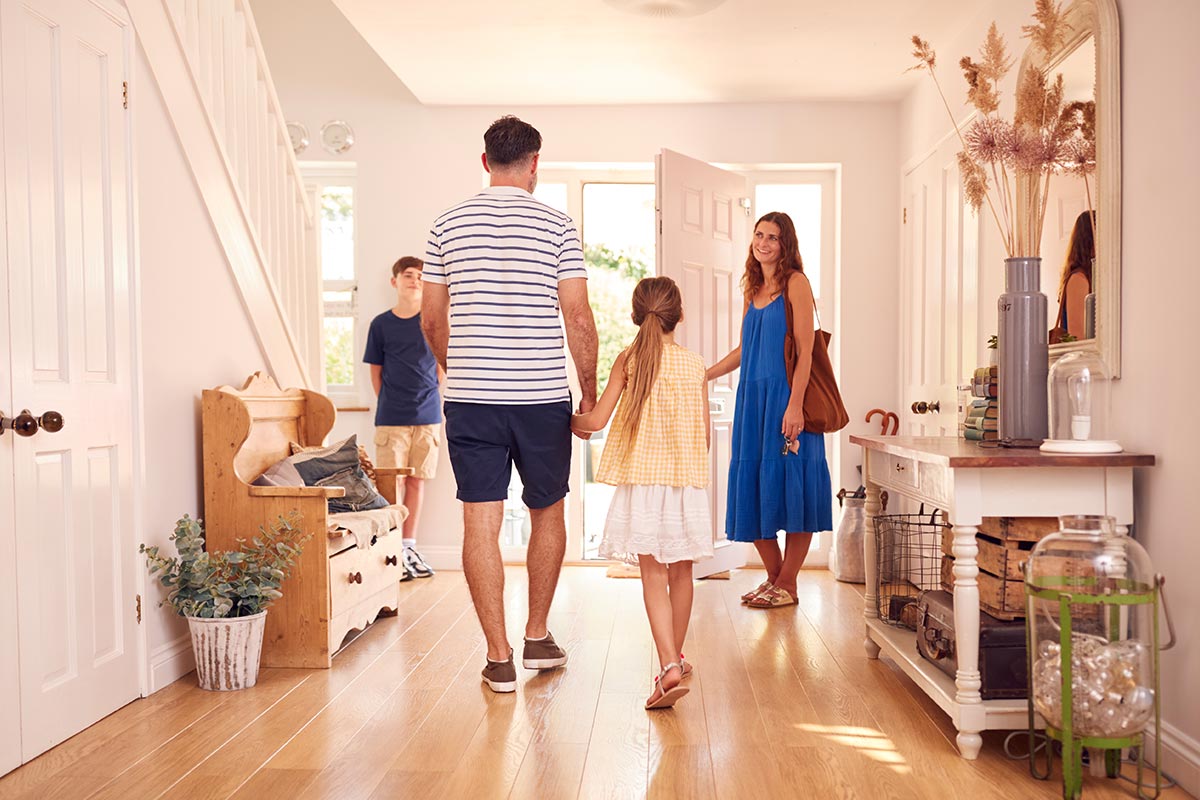 A family of four enters a bright hallway. A man and a girl hold hands, a woman holds a bag by the door, and a boy stands near the stairs.