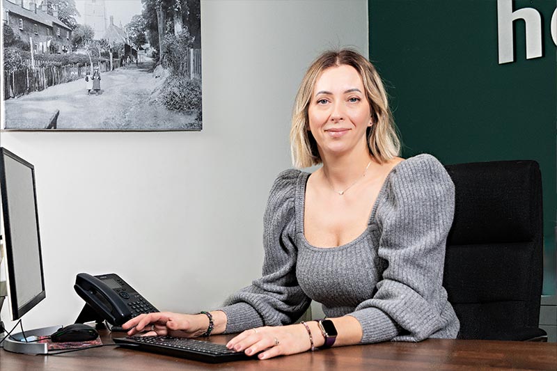 Person seated at a desk, using a computer and phone, with a framed black and white photo on the wall.