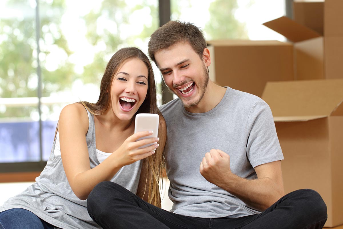 A smiling couple sits on the floor in front of boxes, both wearing gray shirts. They eagerly look at a phone, possibly celebrating good news from Homeworks Estate Agents.